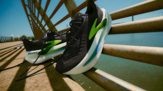 Runner striding on a paved path wearing rockered road shoes; a dashed line illustrates heel-to-toe drop and the upturned toe spring.