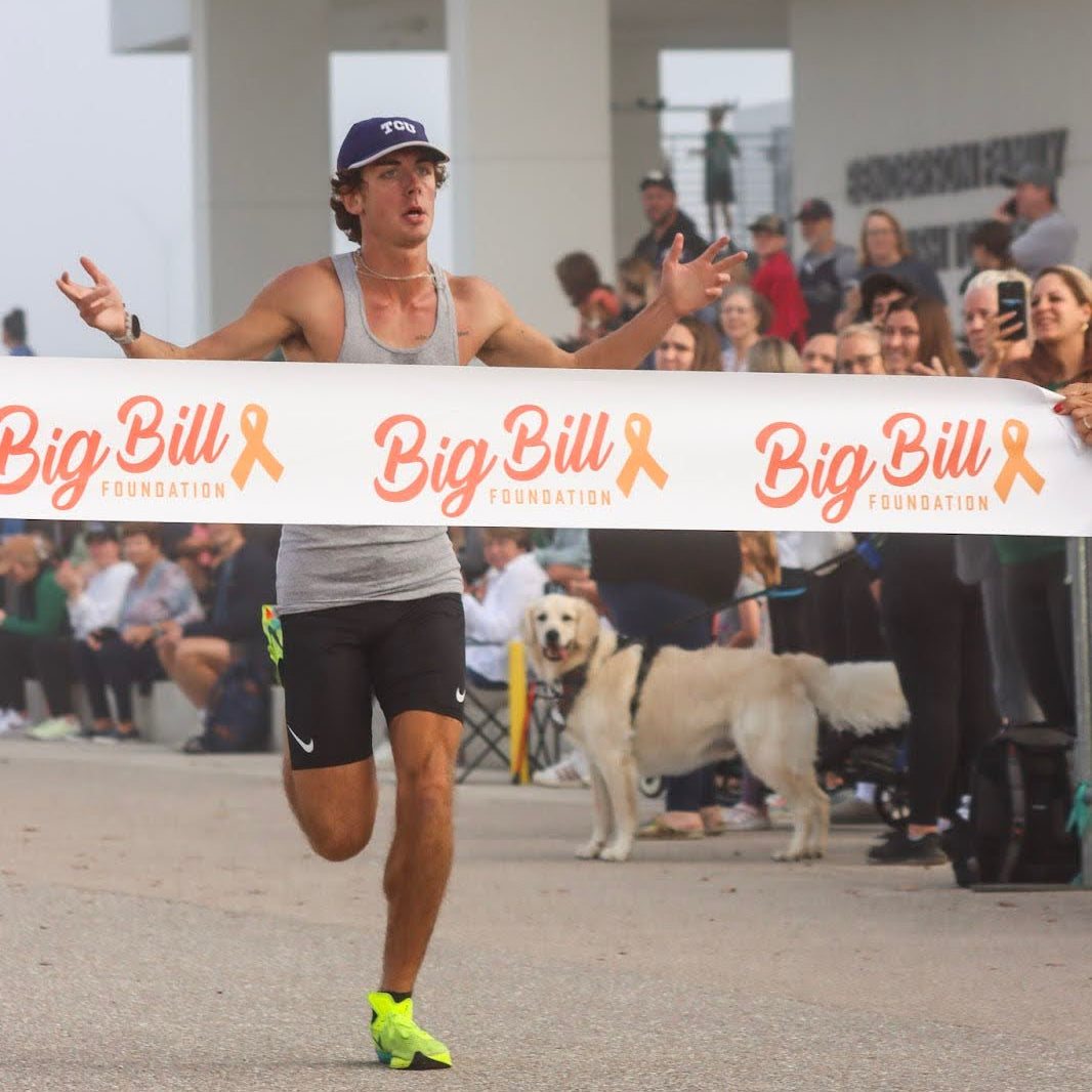 Person crossing a finish line with 'Big Bill' banner, surrounded by spectators and a dog.