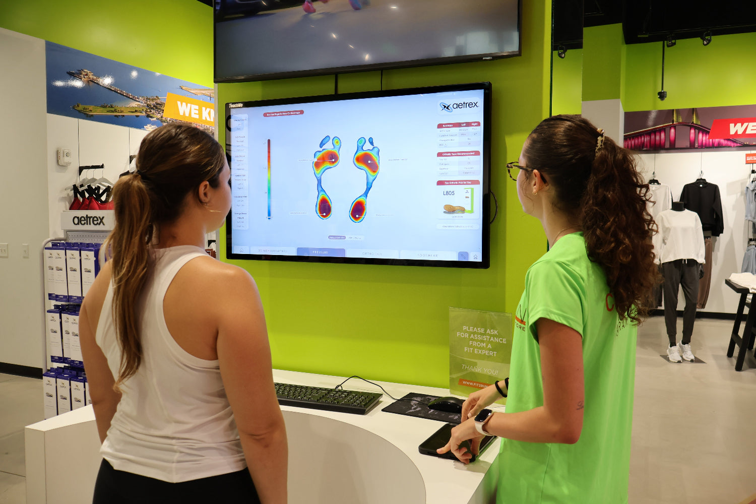Two women interacting with a digital footprint analysis device in a store setting.